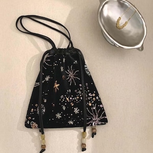 Puede incluir: Bolso negro de terciopelo con cordón y diseños de estrellas y flores con cuentas. El bolso tiene cordones negros y detalles de cuentas en la parte inferior. Un collar de oro descansa en un plato plateado.