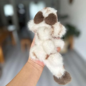 May include: A plush toy in the shape of a dog's paw, featuring a soft, fluffy texture. The toy is primarily white with brown spots and accents, creating a realistic and cuddly appearance. The toy is held in a hand.