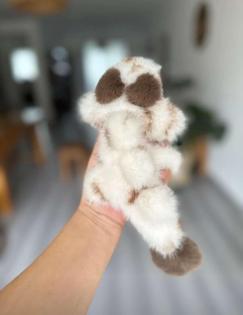 May include: A plush toy in the shape of a dog's paw, featuring a soft, fluffy texture. The toy is primarily white with brown spots and accents, creating a realistic and cuddly appearance. The toy is held in a hand.