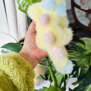 May include: A fluffy, yellow plush toy with light blue and pink accents. The toy has a bow shape at the top and three pink pom-poms. The bottom of the toy has light blue accents. The toy is held in a hand.