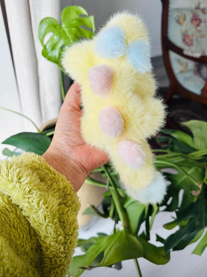 May include: A fluffy, yellow plush toy with light blue and pink accents. The toy has a bow shape at the top and three pink pom-poms. The bottom of the toy has light blue accents. The toy is held in a hand.