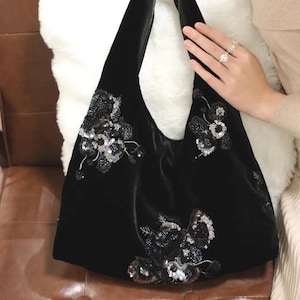 Puede incluir: Bolso tote de terciopelo negro con adornos florales de lentejuelas y cuentas. El bolso tiene una parte superior ancha y abierta y correas largas para el hombro. El diseño floral se concentra en la parte delantera del bolso, con lentejuelas y cuentas plateadas y negras.