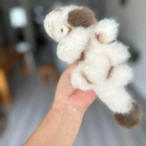 May include: A plush toy dog with a white body and brown spots. The soft toy has a fluffy texture and a small, round head. The dog is held in a hand, with the background blurred.