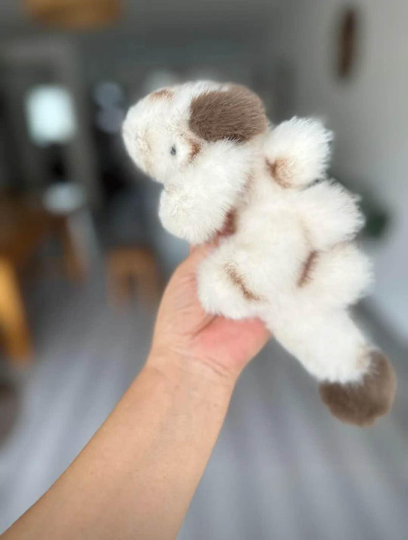 May include: A plush toy dog with a white body and brown spots. The soft toy has a fluffy texture and a small, round head. The dog is held in a hand, with the background blurred.