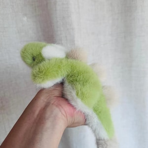 May include: A plush toy dinosaur in shades of green, white, and tan. The soft toy has a fluffy texture and a friendly, cartoonish design. The dinosaur is a light green color with white accents and tan spikes.