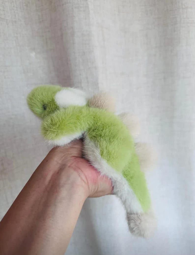 May include: A plush toy dinosaur in shades of green, white, and tan. The soft toy has a fluffy texture and a friendly, cartoonish design. The dinosaur is a light green color with white accents and tan spikes.