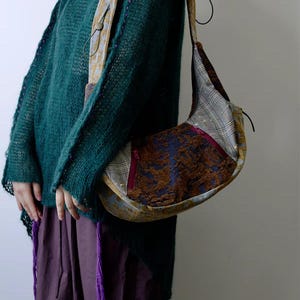 Puede incluir: Un bolso de hombro con forma de media luna, con un diseño de patchwork de varias telas. El bolso incorpora estampados de cuadros, florales y texturizados en marrón y azul. La correa es de color beige claro con un diseño floral. La persona lleva un suéter verde y pantalones morados.
