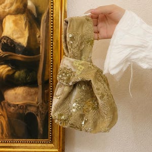 May include: A soft, light olive green velvet knot bag featuring intricate floral embroidery and shimmering gold sequins. The fabric has a plush texture with a distinct sheen, and the design includes a single integrated handle loop for carrying.