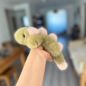 May include: A plush toy in the shape of a green and pink dinosaur. The toy has a soft, furry texture and a rounded head. The dinosaur's back features pink accents, and its feet are also pink.