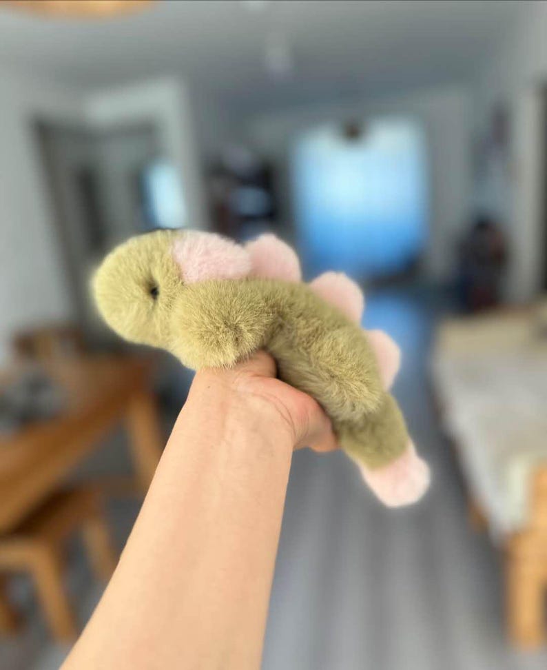 May include: A plush toy in the shape of a green and pink dinosaur. The toy has a soft, furry texture and a rounded head. The dinosaur's back features pink accents, and its feet are also pink.