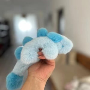 Puede incluir: Un juguete de peluche con forma de dinosaurio, que presenta un cuerpo suave de color azul claro con púas de color azul oscuro a lo largo de la espalda. El juguete tiene una superficie texturizada y difusa, con una forma curva y alargada.
