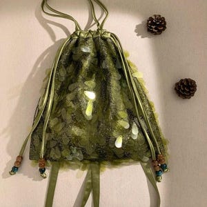 Puede incluir: Un bolso de cordón verde oliva con una capa de malla texturizada y brillante, adornado con numerosas lentejuelas translúcidas en forma de hoja. El bolso incluye correas de cinta de satén a juego y cuentas de madera decorativas en marrón y azul verdoso.