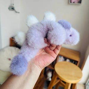 May include: A soft, lavender-coloured toy creature with white accents. The toy has a long body, a rounded head, and small, purple eyes. The creature's fur is fluffy, and it has white, feathery wings. The toy is held in a hand.