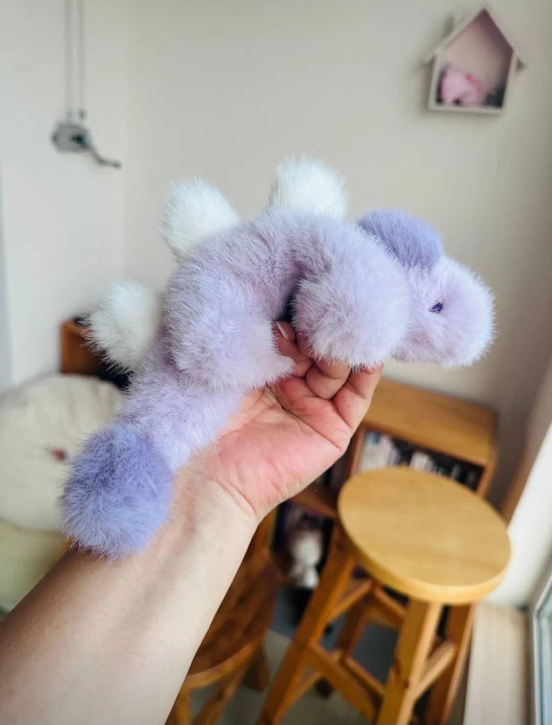 May include: A plush, lavender-colored creature with white accents. The toy has a long body, a rounded head, and small, purple eyes. The creature's fur is soft and fluffy, and it has white, feathery wings. The toy is held in a hand.