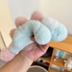 May include: A plush dinosaur toy in pastel colors. The toy features a light blue body with pink and white accents. The soft, furry texture and rounded shape give it a cuddly appearance. The toy is held in a hand.