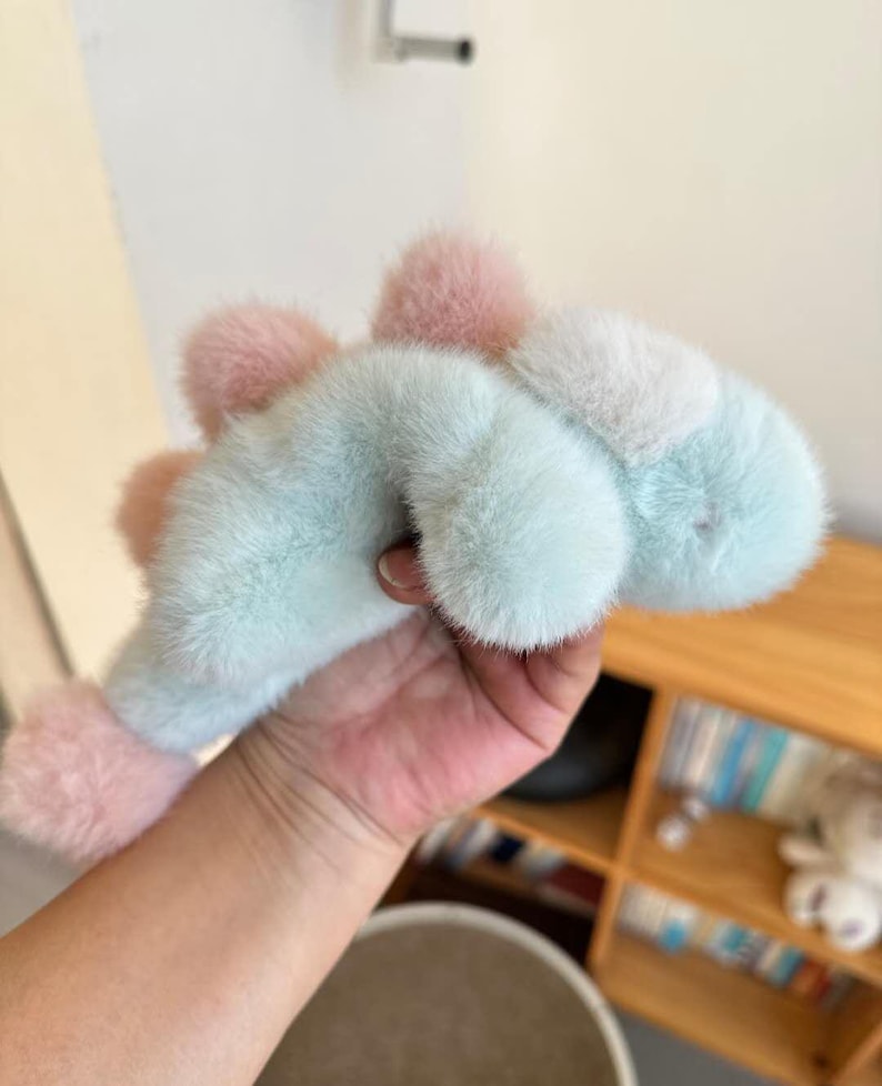 May include: A plush dinosaur toy in pastel colors. The toy features a light blue body with pink and white accents. The soft, furry texture and rounded shape give it a cuddly appearance. The toy is held in a hand.