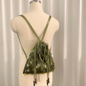 Op de afbeelding: Een saliegroene rugzak met trekkoordsluiting, voorzien van een mesh overlay versierd met bijpassende groene pailletten. De tas heeft dunne schouderbanden en twee lange trekkoorden die eindigen in kleine, veelkleurige houten kralen.