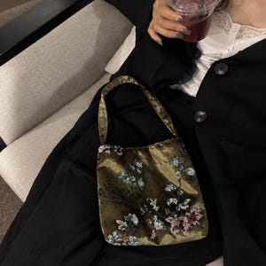 Könnte beinhalten: Eine olivgrüne Samthandtasche mit einem einzelnen Schulterriemen und aufwendiger Blumenstickerei. Das Design umfasst kleine, schimmernde Pailletten in Blau-, Rosa- und Weißtönen, die ein detailliertes botanisches Muster auf der Vorderseite bilden.