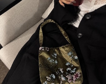 May include: An olive green velvet handbag featuring a single shoulder strap and intricate floral embroidery with blue, pink, and white sequins. The bag has a rectangular shape and a soft, plush texture, showcasing detailed beadwork across the front.