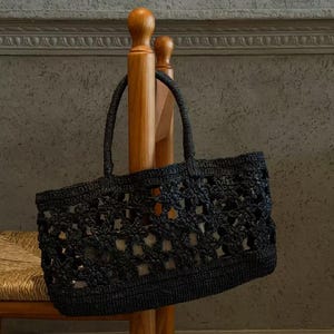 Puede incluir: Un bolso tote negro hecho de material de ganchillo con un patrón floral repetitivo en todo el cuerpo. El bolso incluye dos asas negras a juego y una base sólida y tejida firmemente, lo que proporciona una forma estructurada al accesorio.