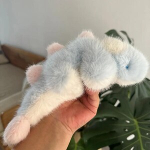 May include: A plush toy in the shape of a sea creature, featuring a soft, furry texture. The toy has a light blue body with white and pink accents. It has a small blue eye and pink fins. The toy is held in a person's hand.