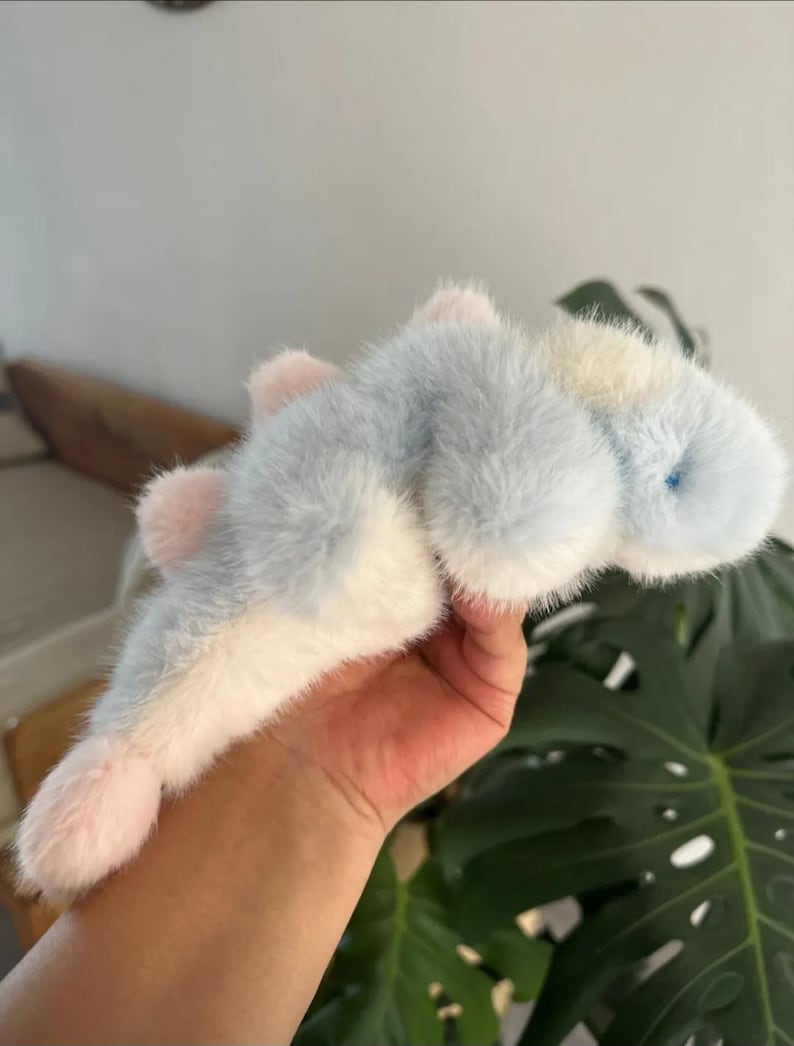 May include: A plush toy in the shape of a sea creature, featuring a soft, furry texture. The toy has a light blue body with white and pink accents. It has a small blue eye and pink fins. The toy is held in a person's hand.