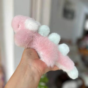 May include: A plush toy in the shape of a pink and white creature with light blue accents. The toy has a soft, furry texture and is held in a person's hand. The toy's design includes rounded shapes and a whimsical appearance.