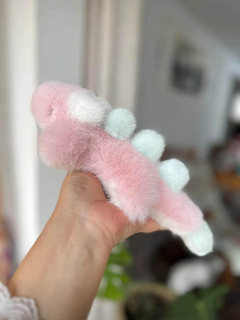 May include: A plush toy in the shape of a pink and white creature with light blue accents. The toy has a soft, furry texture and is held in a person's hand. The toy's design includes rounded shapes and a whimsical appearance.