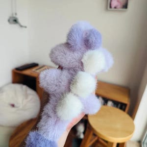 May include: A fluffy, lavender and white accessory with a pom-pom design. The item is held in a hand, and the background includes wooden furniture and a small decorative house.
