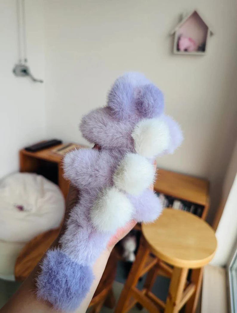 May include: A fluffy, lavender and white accessory with a pom-pom design. The item is held in a hand, and the background includes wooden furniture and a small decorative house.