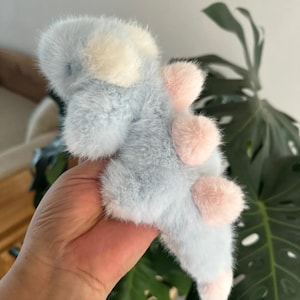May include: A plush toy dinosaur in soft blue, white, and pink. The fluffy toy has a light blue body with white accents on its head and pink spines along its back. The dinosaur's feet are also pink.