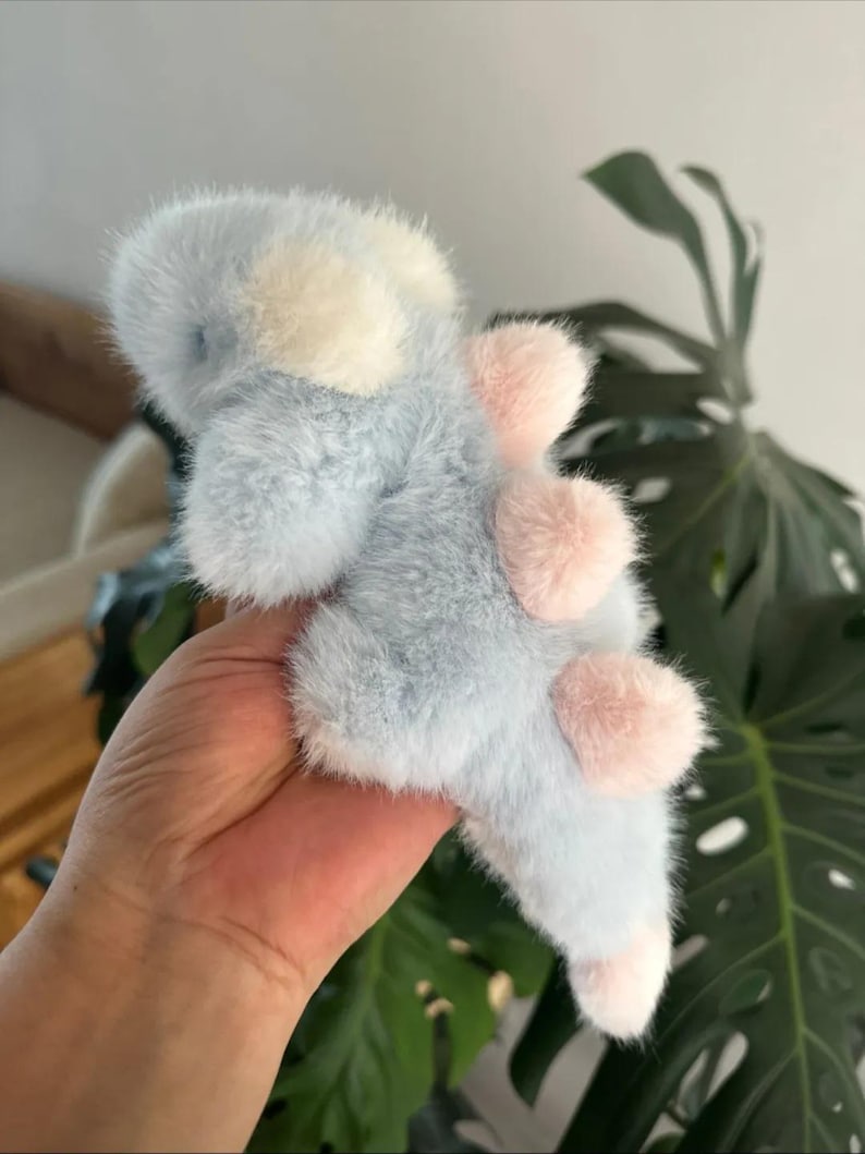 May include: A plush toy dinosaur in soft blue, white, and pink. The fluffy toy has a light blue body with white accents on its head and pink spines along its back. The dinosaur's feet are also pink.