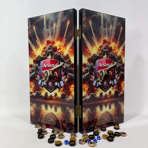 May include: A folding backgammon board featuring the Arsenal football club logo with a cannon and explosion graphic. The board is black with gold-toned metal hinges and is displayed with scattered black and wood-toned checkers and blue dice.