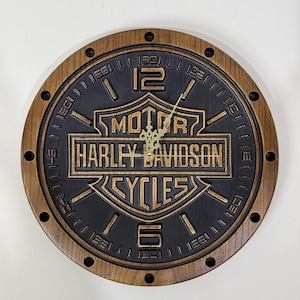 Könnte beinhalten: Runde Holzuhr mit Harley-Davidson-Logo in der Mitte. Das Zifferblatt ist schwarz mit goldfarbenen Zeigern und Ziffern. Der äußere Ring ist braun mit kleinen Löchern. Durchmesser ca. 30 cm.