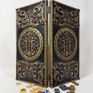 May include: A black and gold backgammon set with ornate carvings and a central medallion design. The set is open, revealing the playing surface, and includes wooden game pieces and blue dice.