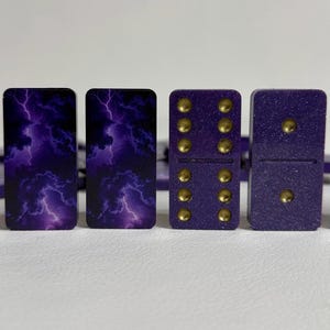 May include: A set of purple dominoes with gold dots. The backs of the dominoes feature a purple and black lightning storm design. The dominoes are arranged on a white surface.