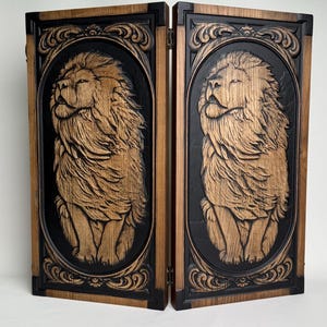 May include: A two-panel wooden screen featuring carved lion portraits. Each panel has a black background with a detailed, light brown lion carving. The frames are dark brown with decorative scrollwork.