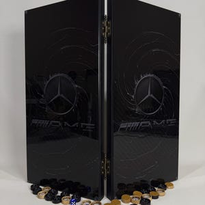 Könnte beinhalten: Ein schwarzer, zweiteiliger Faltbildschirm mit dem Mercedes-Benz AMG-Logo und -Text. Der Bildschirm ist mit messingfarbener Hardware versehen. Eine Sammlung von Spielsteinen, darunter Würfel und Dame, befindet sich am Boden.