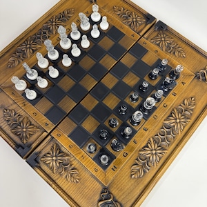 Op de afbeelding: Een houten schaakspel met een gedetailleerde, gesneden rand. Het schaakbord is zwart en bruin, met doorzichtige en witte schaakstukken die zijn opgesteld om te spelen. De set wordt gepresenteerd op een wit oppervlak.