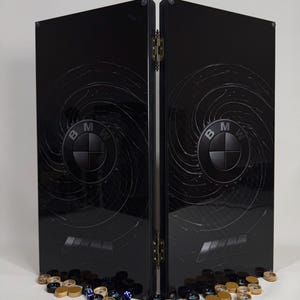 Könnte beinhalten: Ein schwarzes Backgammon-Brett mit dem BMW-Logo und einem Wirbeldesign. Das Brett ist geöffnet und zeigt die Spielfläche. Würfel und Spielsteine in Gold, Schwarz und Blau sind an der Basis verstreut.