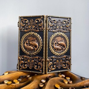 May include: A black and gold backgammon board with detailed carvings. The board is split into two hinged sections, each with a circular emblem depicting fish. Decorative elements and game pieces are present.