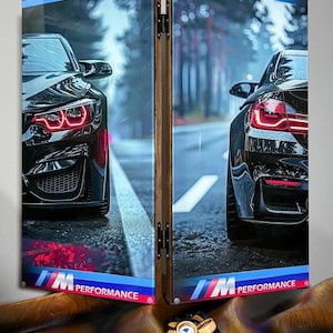 May include: Two-panel screen with images of a black BMW car. The panels have a wooden frame and a design with the BMW logo and the text "M PERFORMANCE". The car's headlights and taillights are illuminated.