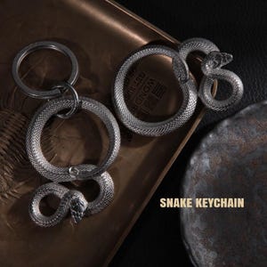 Könnte beinhalten: Silberfarbene Schlangen-Schlüsselanhänger. Die Schlüsselanhänger haben die Form einer zusammengerollten Schlange mit detaillierten Schuppen und einem Schlangenkopf. Der Text "SNAKE KEYCHAIN" befindet sich unten rechts. Die Schlüsselanhänger liegen auf einem braunen Tablett.