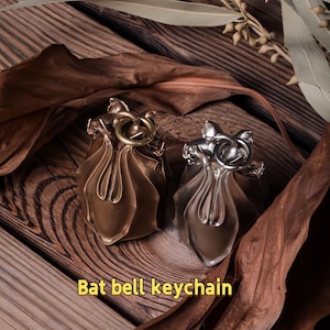 May include: Two bat bell keychains, one bronze and one silver, rest on a wooden surface. The keychains have a bell shape with a bat-like design at the top and a ring for attaching keys. The text "Bat bell keychain" is at the bottom.