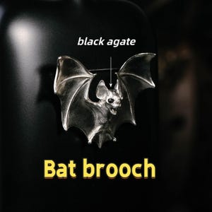 May include: A silver bat brooch with detailed wings and facial features. The text "black agate" and "Bat brooch" are displayed on a black background. The brooch has a pin closure.