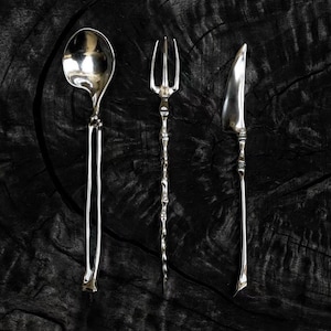 May include: A set of three silver-toned utensils consisting of a spoon, a three-pronged fork, and a knife. Each piece features a slender, textured handle with an organic, bone-like appearance. The text "ingo studio" is printed at the bottom.