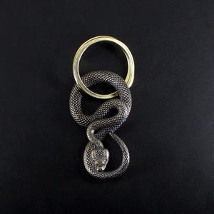 Puede incluir: Un llavero de serpiente metálica detallada, con un anillo dorado. La serpiente está enrollada, con escamas texturizadas y una cabeza detallada. El fondo es negro liso.