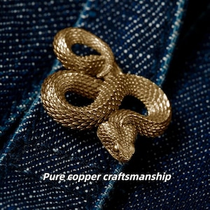 Op de afbeelding: Een gedetailleerde, goudkleurige slangvormige broche. De broche is gemaakt van koper en heeft een getextureerd, geschubd oppervlak. De tekst "Pure copper craftsmanship" wordt eronder weergegeven.