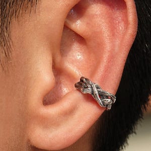 May include: A silver-toned ear cuff featuring a textured, braided metal design that wraps around the concha of the ear. The metal has a darkened, oxidised finish in the recessed areas to highlight the intricate, woven rope-like pattern.