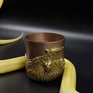 May include: A copper-coloured tumbler with a textured surface and a decorative brass snake scale design wrapped around the base. The tumbler is set against a black background with a yellow snake.